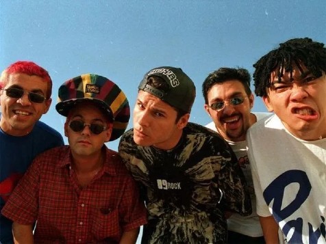 A imagem apresenta o grupo musical Mamonas Assassinas, em um médio plano com o céu de fundo, há cinco pessoas de frente fazendo caretas, típico da personalidade do grupo
